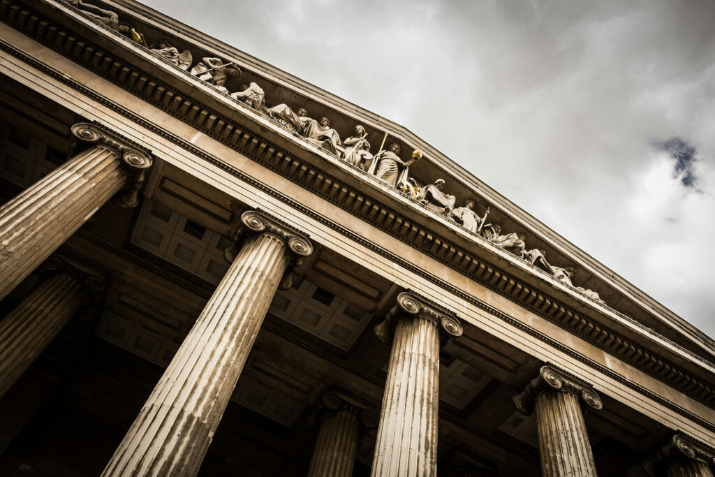 Façade d'un bâtiment de justice avec des colonnes imposantes et des sculptures en relief sur le fronton sous un ciel nuageux, symbolisant la grandeur et la solennité du droit.